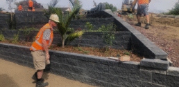 man in a hard hat and a high vis vest working on a retaining wall with a digger in partial view, and other men in the background